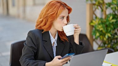 Genç kızıl saçlı iş kadını akıllı telefonla selfie çekiyor. Kahve dükkanının terasında kahve içiyor.