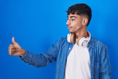 Young hispanic man standing over blue background looking proud, smiling doing thumbs up gesture to the side 