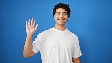 Genç latin adam, mavi arka planda gülümseyerek merhaba diyor.