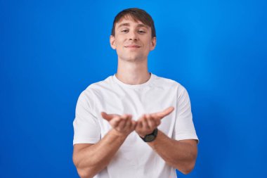 Mavi arka planda dikilen beyaz, sarışın bir adam avuçları birbirine gülümseyen ve el hareketi yapan bir adam. Bekleme ve koruma 