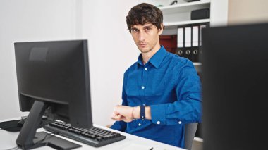 Genç İspanyol iş adamı bilgisayar kullanıyor. Ofiste ciddi yüzlü bir saat kullanıyor.