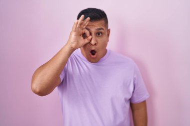 Pembe arka planda duran genç İspanyol adam şaşırmış yüz ifadesiyle şaşırmış, parmaklarının arasından bakan bir jest yapıyor. İnançsız ifade. 