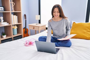 Young beautiful hispanic woman student reading book studying on bed at bedroom