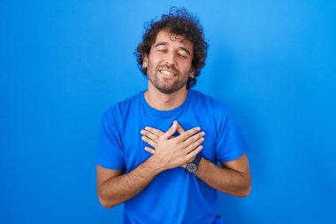 Mavi arka planda duran İspanyol genç adam elleri göğsünde, gözleri kapalı ve yüzünde minnet dolu bir jest ile gülümsüyor. sağlık kavramı. 