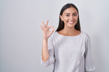 Young hispanic woman standing over white background smiling positive doing ok sign with hand and fingers. successful expression. 