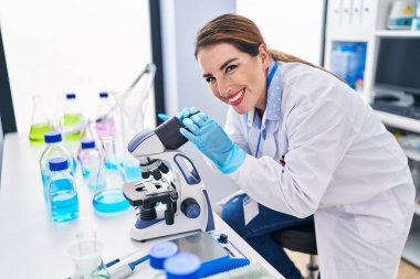 Young beautiful hispanic woman scientist smiling confident using microscope at laboratory