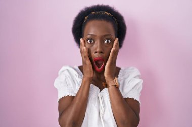 Pembe arka planda duran kıvırcık saçlı Afrikalı kadın. Korkmuş ve şok olmuş, şaşırmış ve hayrete düşmüş. Yüzünde elleri olan. 