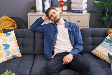 Kafkasyalı genç adam, evinde kanepede, elleri başının üstünde rahatlamış bir şekilde oturuyor.