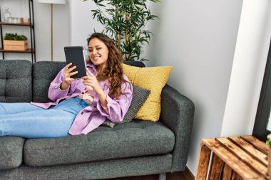 Evdeki kanepede uzanmış dokunmatik ped kullanan genç, güzel İspanyol kadın.