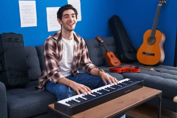 Young hispanic man playing piano at music studio smiling and laughing hard out loud because funny crazy joke.