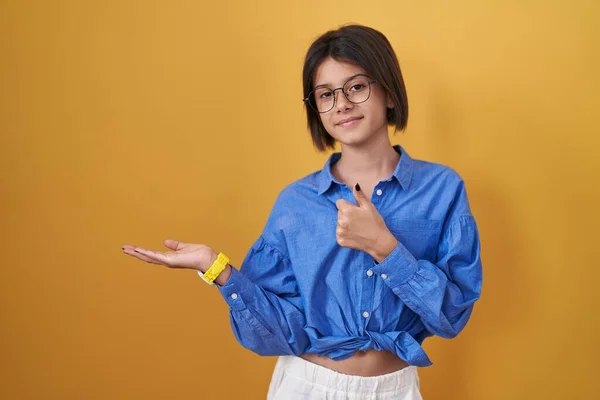 stock image Young girl standing over yellow background showing palm hand and doing ok gesture with thumbs up, smiling happy and cheerful 