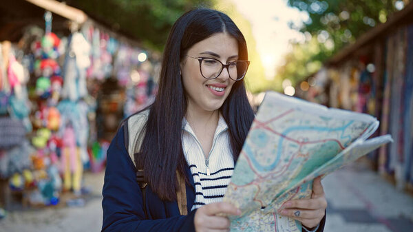 Young hispanic woman tourist wearing backpack looking at city map at street market