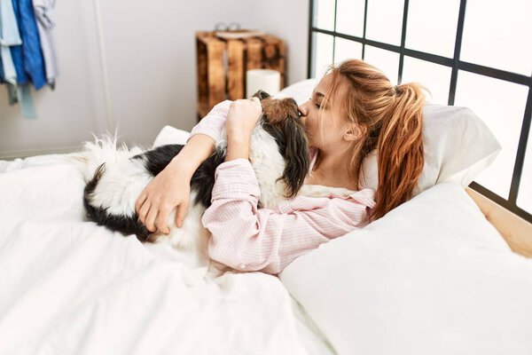 Молодая белая женщина лежит на кровати обнимая и целуя собаку в спальне