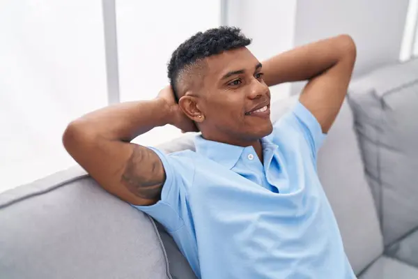 Young latin man relaxed with hands on head sitting on sofa at home