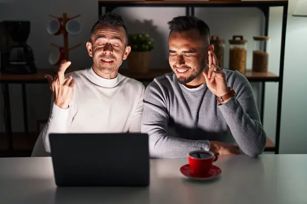 Homosexual couple using computer laptop gesturing finger crossed smiling with hope and eyes closed. luck and superstitious concept.