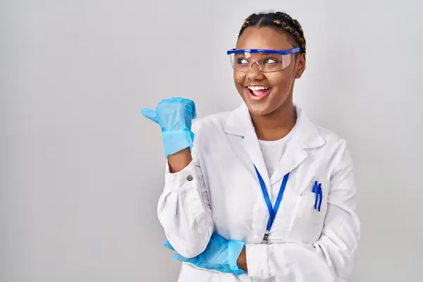 身穿科学家长袍 头戴辫子的非洲裔美国女人 面带微笑 面带微笑 并竖起大拇指指向旁边 — 图库照片