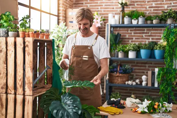 Young blond man florist watering plant working at flower shop