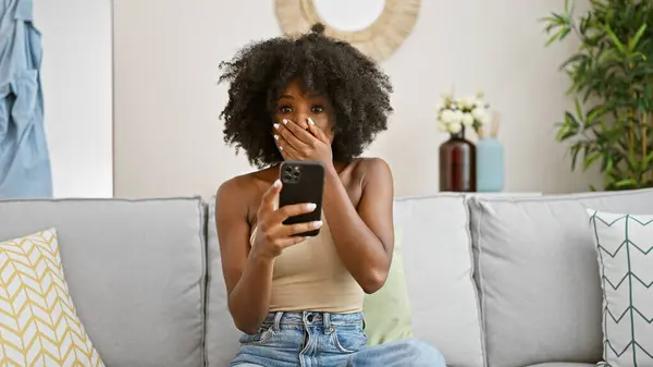 Afroamerykanka Pomocą Smartfona Siedzi Kanapie Zaskoczony Domu — Zdjęcie stockowe