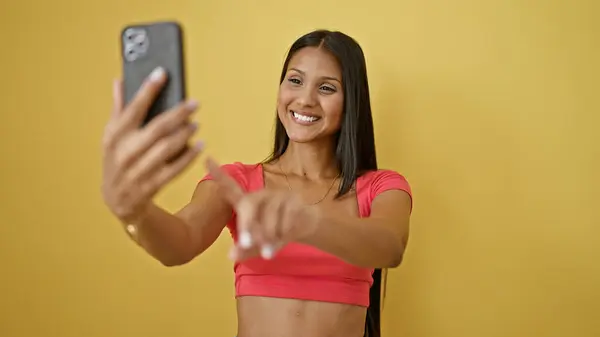 年轻的拉丁女人笑着自信地用智能手机在孤立的黄色背景下自拍 — 图库照片