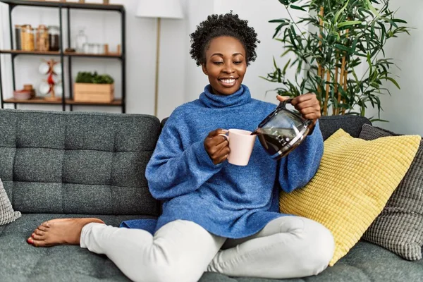 Afro-Amerikalı kadın evdeki kanepede oturan fincana kahve dolduruyor.