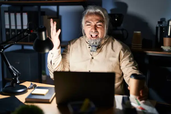 Middle age man with grey hair working at the office at night crazy and mad shouting and yelling with aggressive expression and arms raised. frustration concept.