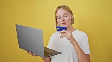 Laptop ve kredi kartıyla alışveriş yapan genç sarışın kadın izole edilmiş sarı arka planda