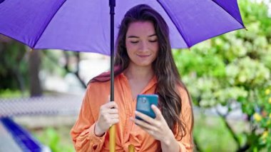 Parkta akıllı telefon kullanan güzel İspanyol bir kadın şemsiye tutuyor.
