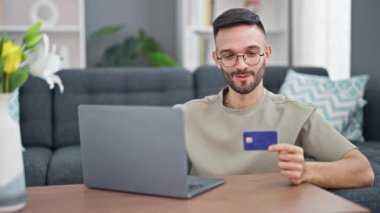 Genç İspanyol adam evde dizüstü bilgisayar ve kredi kartıyla alışveriş yapıyor.