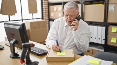 Middle age grey-haired man ecommerce business worker talking on smartphone writing on package at office