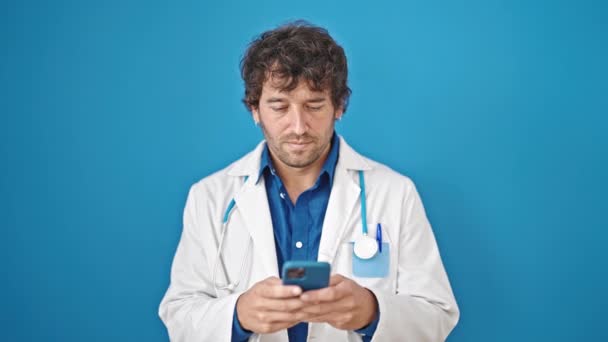 Молодий Іспаномовний Лікар Використовує Смартфон Посміхаючись Над Ізольованим Синім Тлом — стокове відео