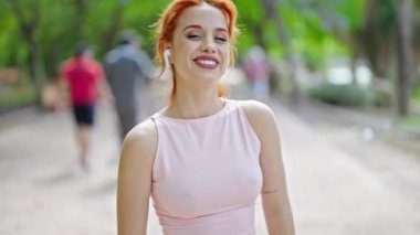 Spor giyim ve kulaklık takan genç kızıl saçlı kadın parkta gülümsüyor.