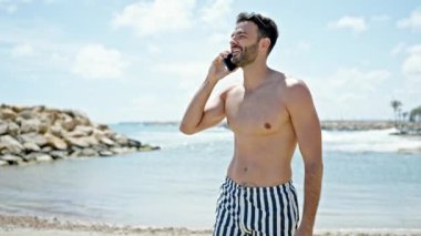 Genç İspanyol turist mayo giyiyor. Akıllı telefondan konuşuyor. Kumsalda gülümsüyor.