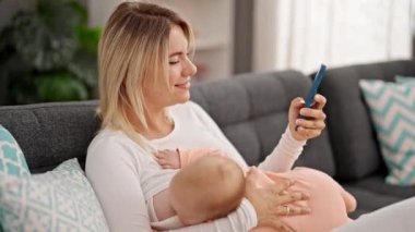 Anne ve kız kanepede oturup bebeği emzirmek için akıllı telefon kullanıyorlar.