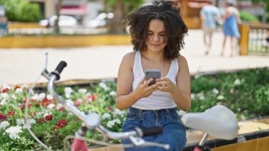 Genç ve güzel İspanyol bir motorcu parkta bankta otururken akıllı telefonuyla selfie çekiyor.