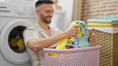 Çamaşır odasında çamaşır makinesine yaslanan elbiseli İspanyol bir adam sepeti tutuyor.