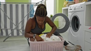 Çamaşır odasında çamaşır sepetinde kıyafet arayan Afrikalı Amerikalı bir kadın.