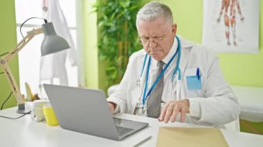 Klinikte rapor yazmak için dizüstü bilgisayar kullanan orta yaşlı, kır saçlı bir adam.