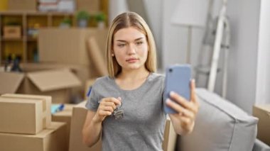 Genç sarışın kadın yeni evinde elinde anahtarla akıllı telefondan selfie çekiyor.