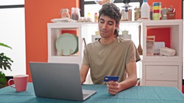 Laptop ve kredi kartıyla alışveriş yapan İspanyol genç adam yemek odasında masada oturuyor.