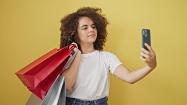 Güzel İspanyol bir kadın alışveriş poşetleri tutuyor ve izole edilmiş sarı arka planda akıllı telefondan selfie çekiyor.