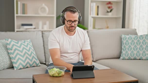 Grå Håret Mand Spiser Salat Ved Hjælp Touchpad Hovedtelefoner Derhjemme – Stock-video