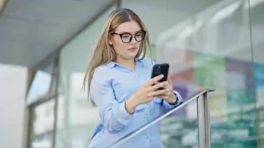 Sarışın genç bir iş kadını sokakta ciddi bir ifadeyle akıllı telefon kullanıyor.