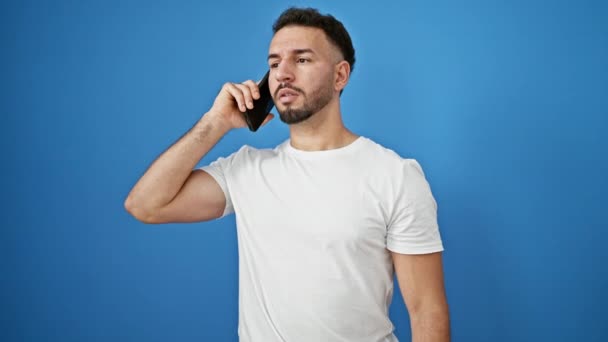 年轻的Arab男子在智能手机上说话 看着手表在孤立的蓝色背景下离去 — 图库视频影像