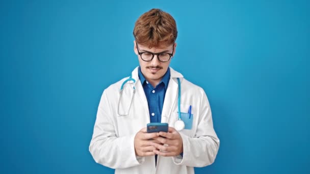 年轻的惊慌失措的医生用智能手机对着孤立的蓝色背景微笑 — 图库视频影像