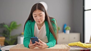 Genç Çinli kadın akıllı telefon kullanıyor. Çamaşır odasındaki ütü masasına yaslanıyor.