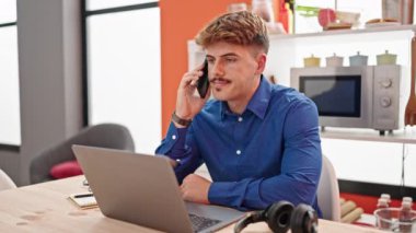 Genç İspanyol adam yemek odasında dizüstü bilgisayar kullanarak akıllı telefondan konuşuyor.