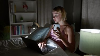 Akıllı telefon ve dizüstü bilgisayar kullanan genç sarışın kadın yatak odasında oturuyor.