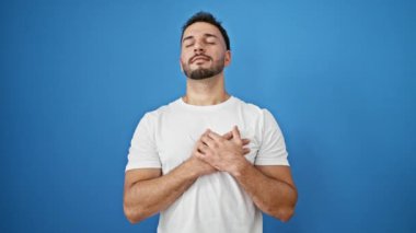 Genç Arap adam, elleri kalbimde, mavi arka planda, kendinden emin gülümsüyor.