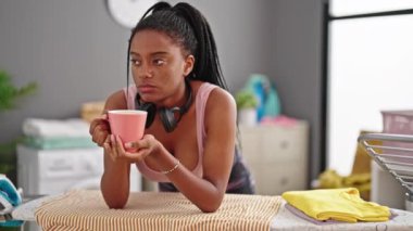 Afrika kökenli Amerikalı kadın kahve içiyor. Çamaşır odasında ütü masasına yaslanıyor.