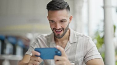 Latin asıllı genç adam, kahvecinin terasında video oyunu oynarken kendine güveniyor.